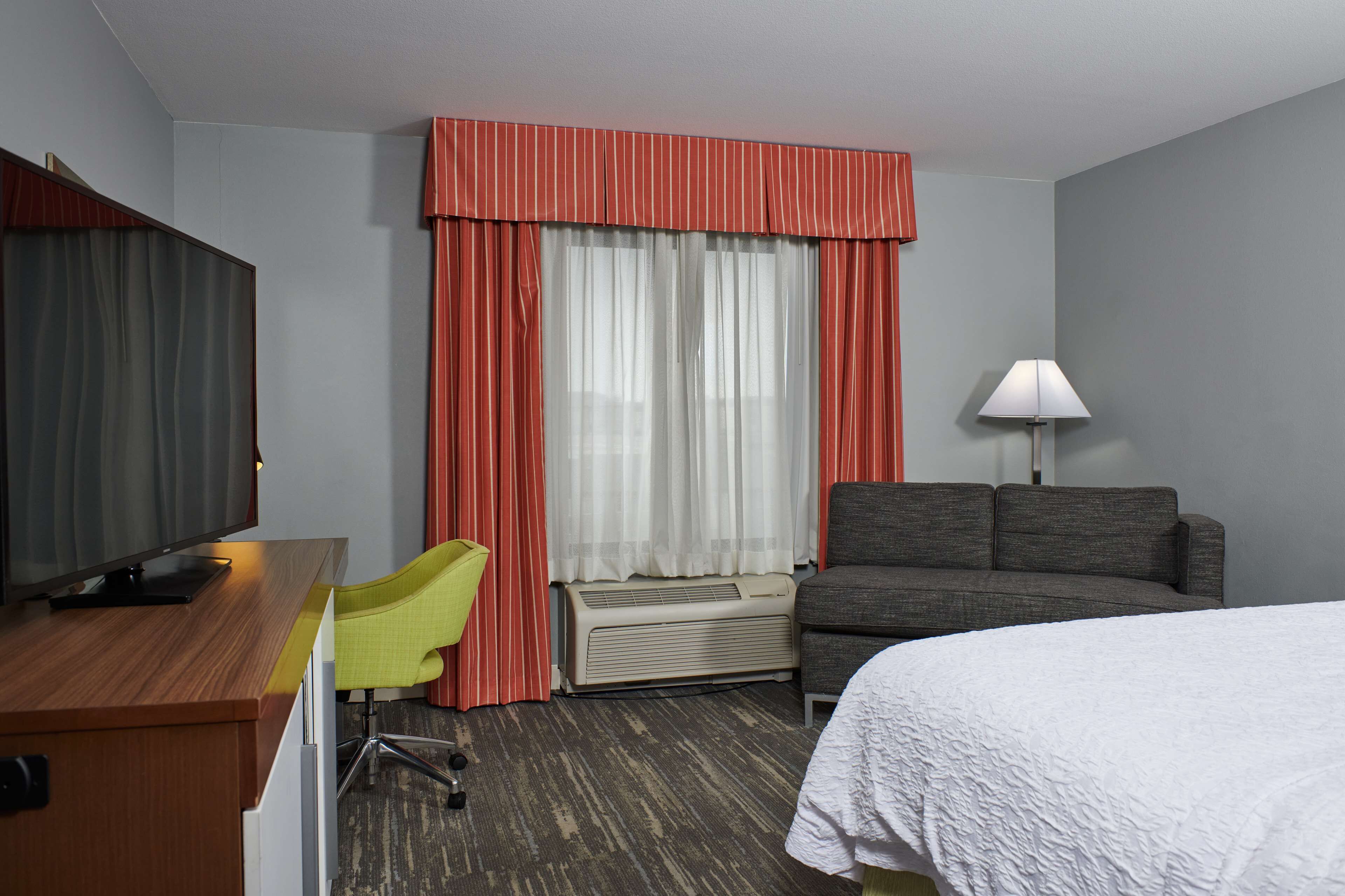 Habitación de hotel con cama, escritorio con televisor, sillón, sofá y ventana con cortinas.