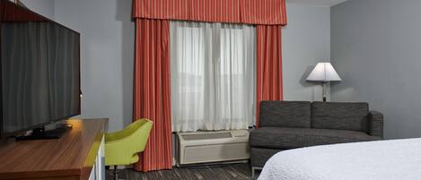 Habitación de hotel con cama, escritorio con televisor, sillón, sofá y ventana con cortinas.