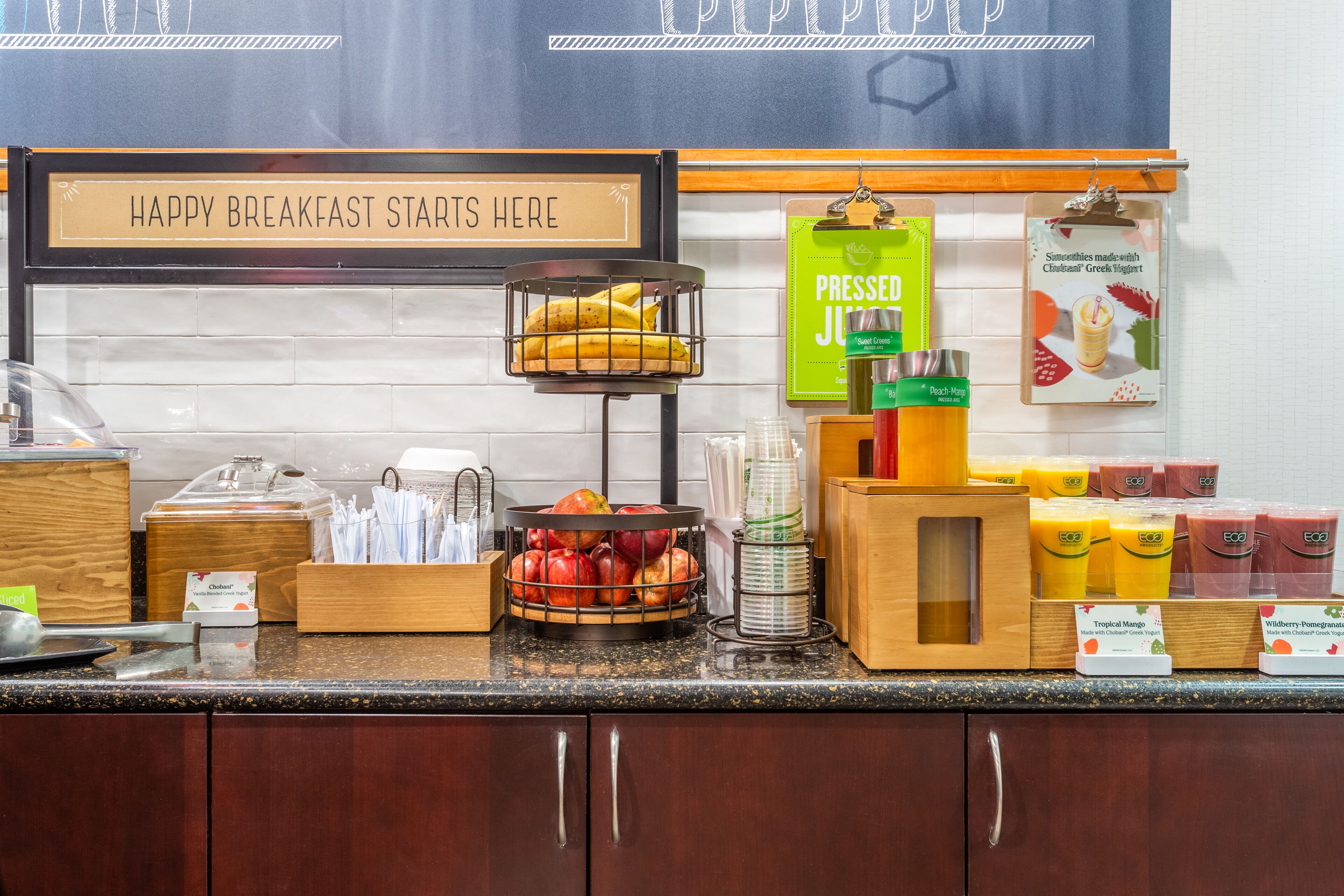 Una barra de desayuno con un cartel que dice "HAPPY BREAKFAST STARTS HERE", una cesta de frutas, dispensadores de jugo y un tablero de menú.