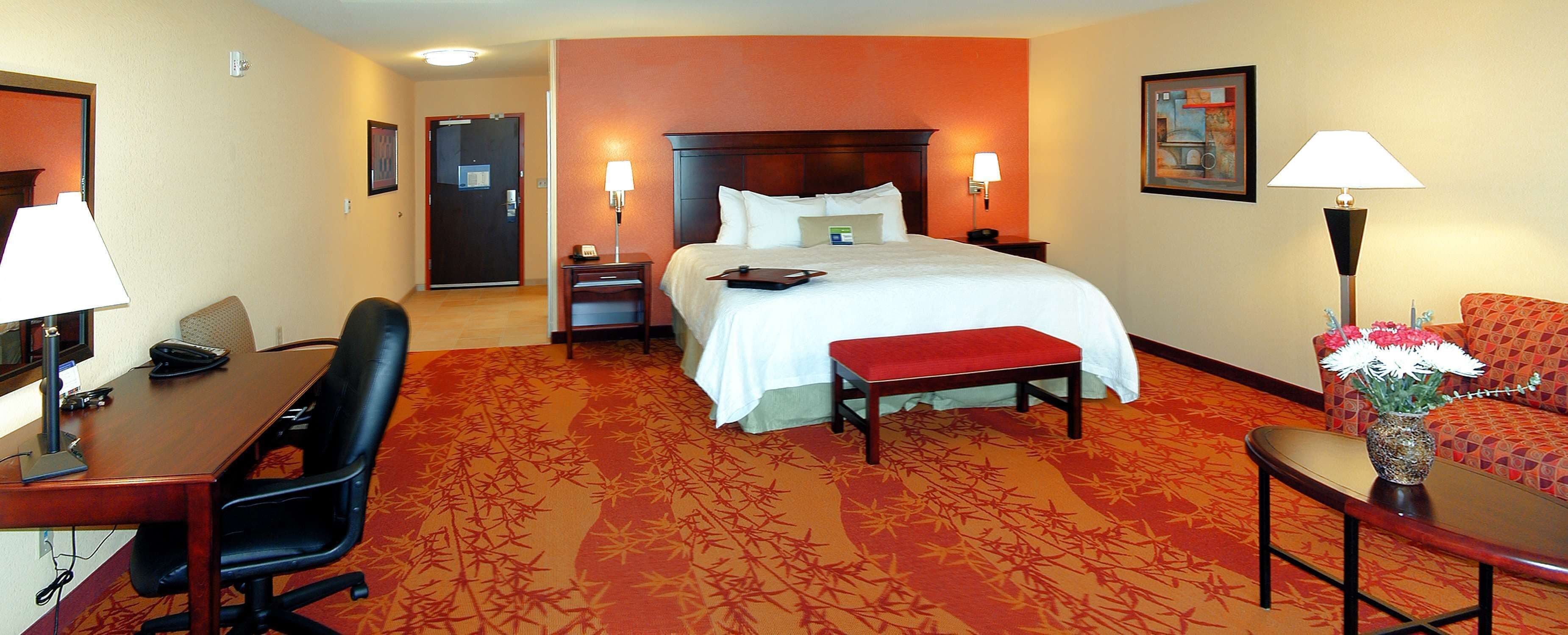 A hotel room with a bed, bedside table, lamp, desk, chair, and a small table with flowers.