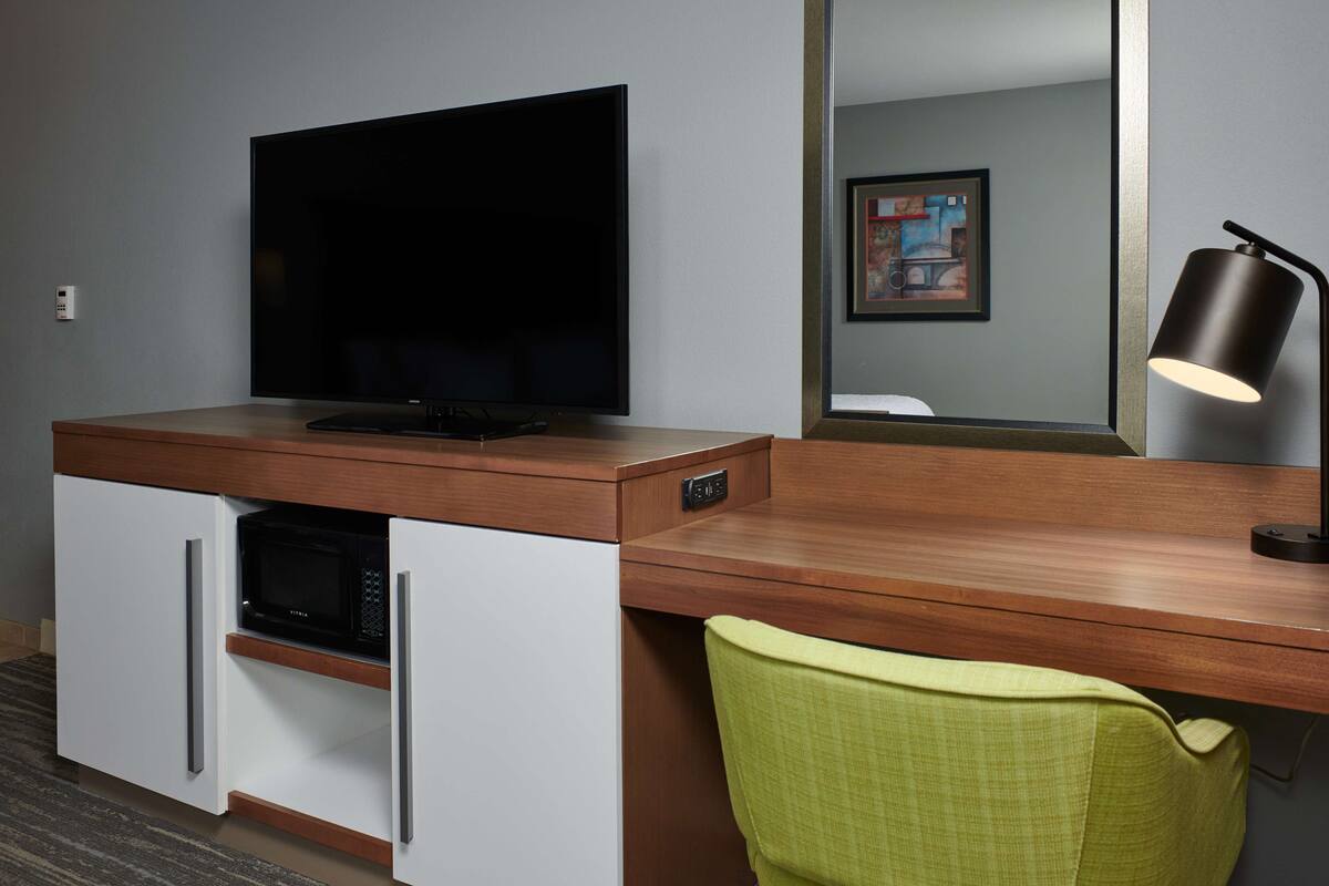 A hotel room with a wooden desk, a flat-screen TV, a microwave, a green chair, and a lamp.