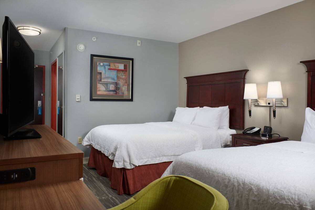 A hotel room with two beds, a TV, a painting on the wall, and a desk with a lamp.
