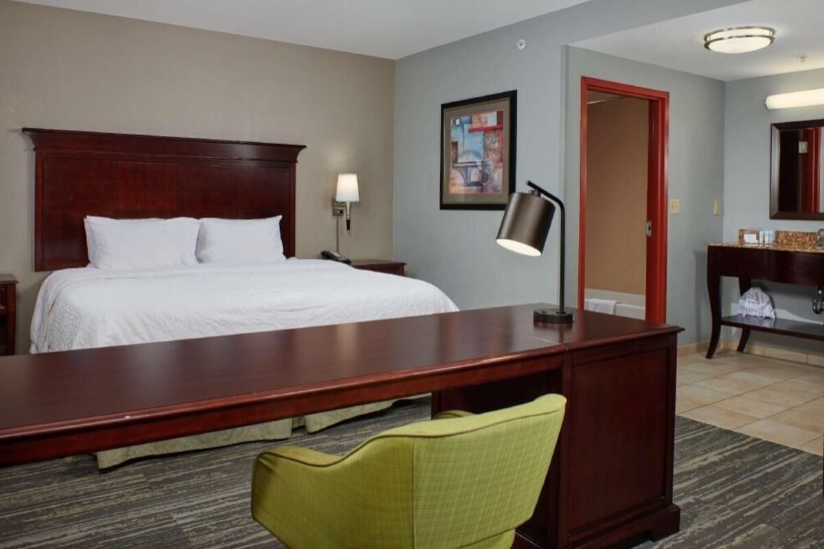 A hotel room with a large bed, a desk, a chair, a lamp, a mirror, and a door.