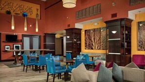 A hotel lobby with a dining area, blue chairs, a sofa with cushions, and a television on the wall.