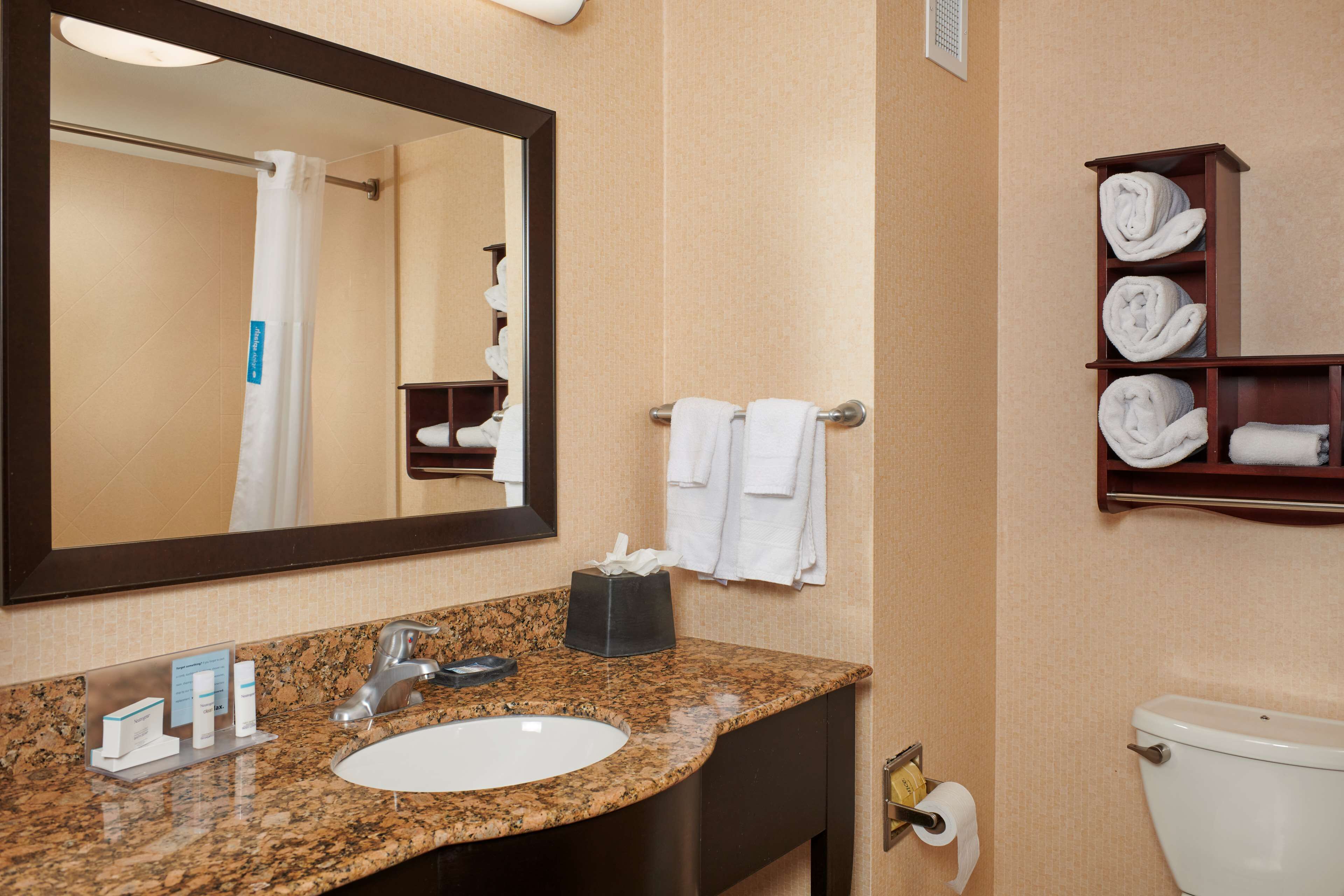 A bathroom with a granite countertop sink, a mirror, a towel rack with white towels, a shelf with neatly folded towels, and a toilet.