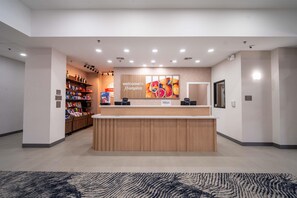 Un espace d’accueil d’hôtel avec un comptoir en bois, une enseigne « Bienvenue à Hampton » et une décoration murale sur le thème des fruits.