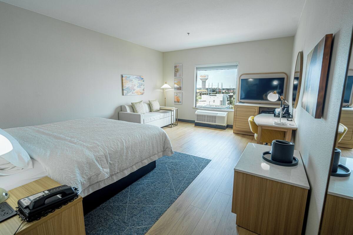 A hotel room with a bed, desk, chair, lamp, and a large window.