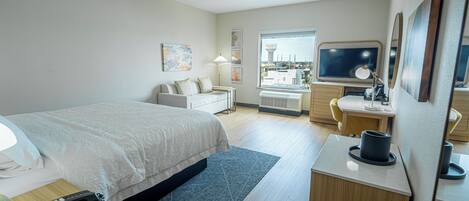 A hotel room with a bed, desk, chair, lamp, and a large window.