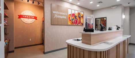 A hotel reception area with a counter, a wall sign reading 'Welcome to Hampton,' and a sign for 'Treats' with snacks and sundries.