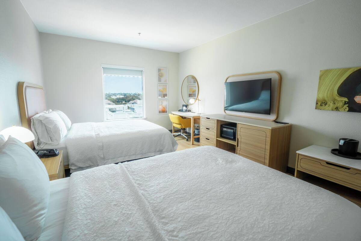 A hotel room with two beds, a desk with a computer, a TV, and a window with a city view.