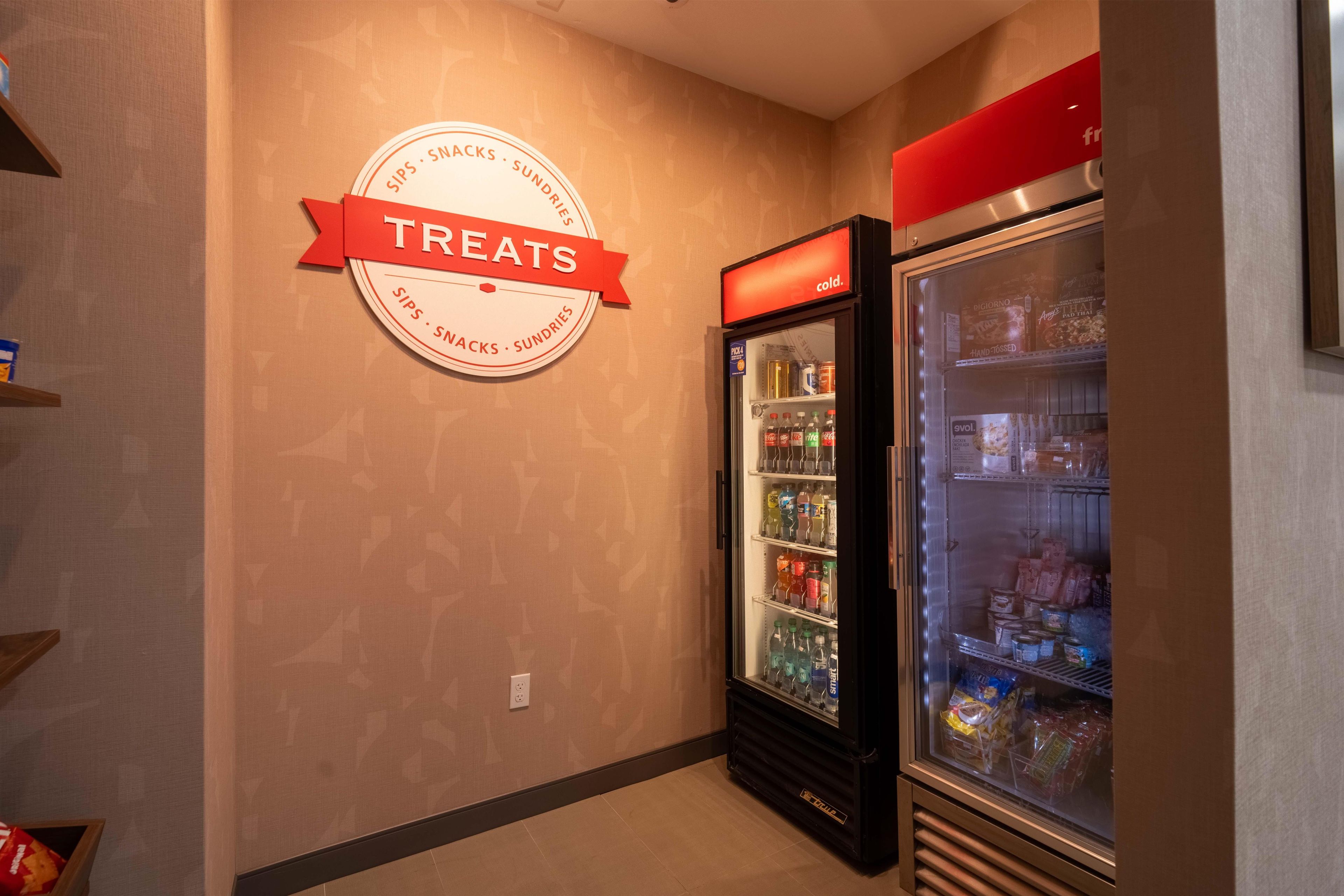 A wall with a circular sign indicating a treats section, a refrigerator stocked with beverages and snacks, and a shelf with packaged items.