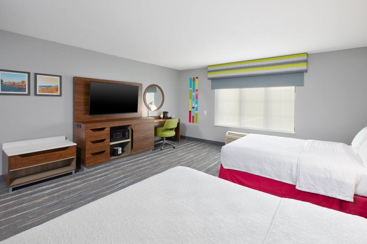 A hotel room with a TV, a desk with a chair, two beds, a window with a view, and a wall-mounted mirror.