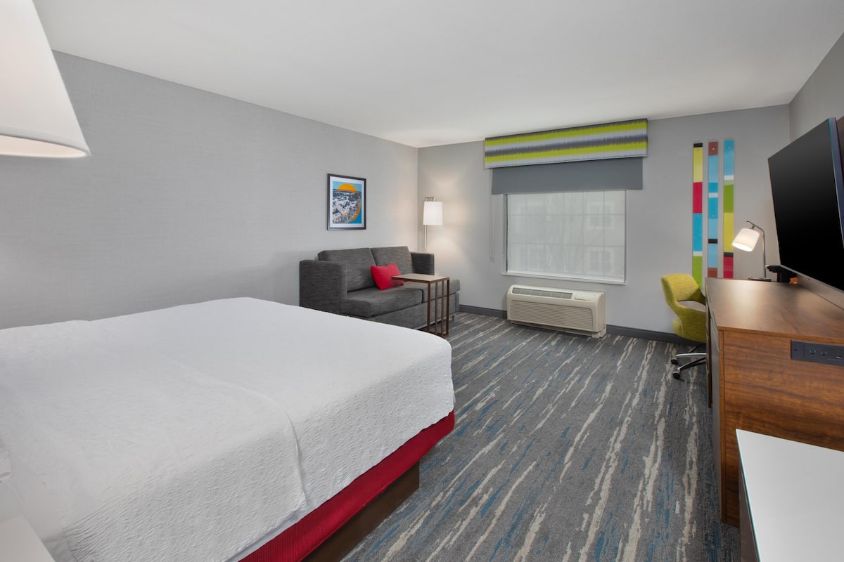 A hotel room with a large bed, a desk, a chair, a TV, a window, and a painting on the wall.
