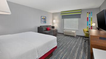 A hotel room with a large bed, a desk, a chair, a TV, a window, and a wall-mounted air conditioner.