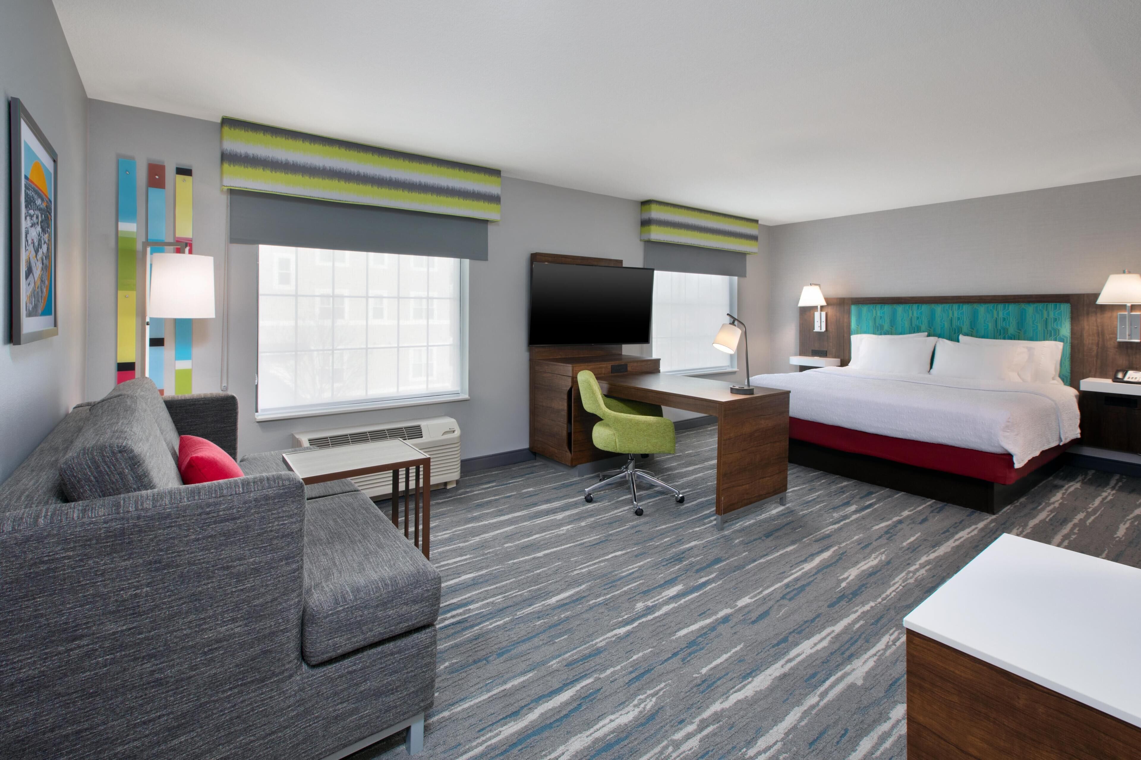 A hotel room with a bed, a desk with a chair, a TV, a window, and a sofa.