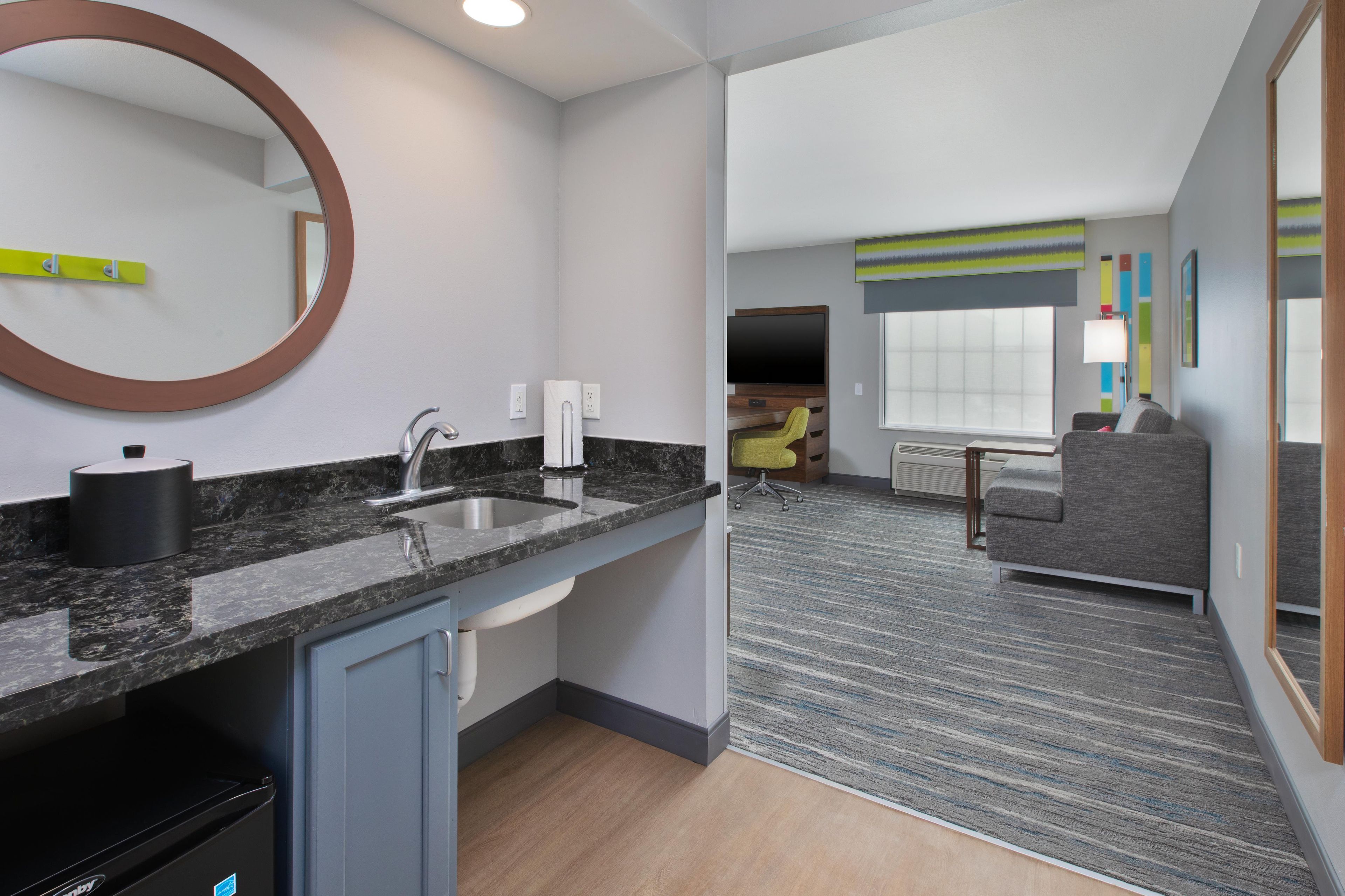A modern hotel room with a kitchen area, a seating area with a sofa and armchair, and a large window.