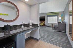 A modern hotel room with a kitchen area, a seating area with a sofa and armchair, and a large window.