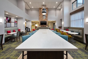 A modern hotel lobby with a long communal table, seating areas, and a flat-screen TV mounted on a stone wall. - Hampton Inn & Suites St. Cloud, MN (St Cloud)