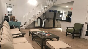 Amplia sala de estar con un conjunto de sofás, una mesa baja con revistas, un sillón verde y una escalera con barandilla.