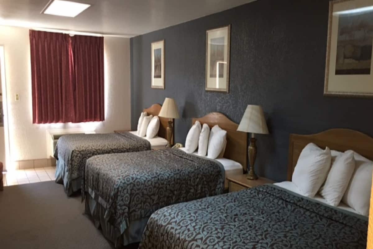 Ett hotellrum med tre sängar, alla med mönstrat sänglinne och sänggavlar. Det finns två sängbord med lampor, ett fönster med röda gardiner och en inramad tavla på väggen.