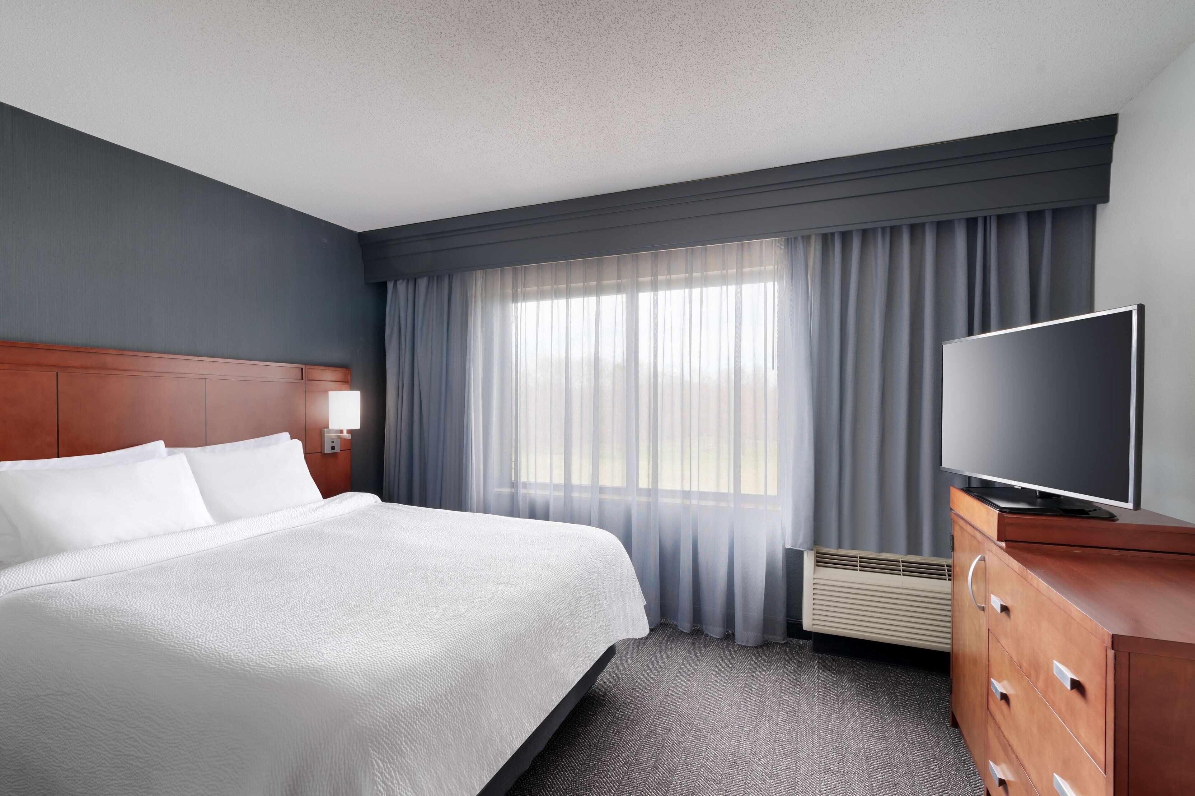 A hotel room with a large bed, a flat-screen TV, a wooden dresser, and a window with sheer curtains.