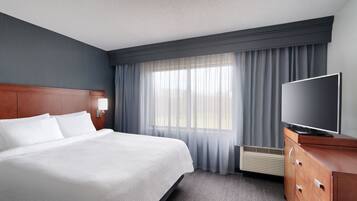 A hotel room with a large bed, a flat-screen TV, a wooden dresser, and a window with sheer curtains.