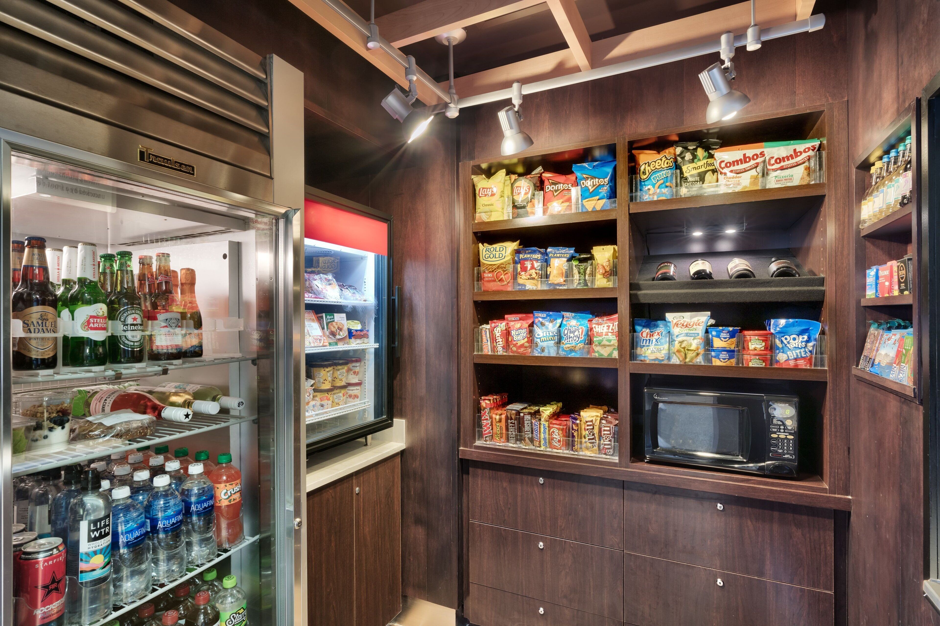 A compact kitchenette with a refrigerator stocked with beverages and snacks, a microwave, and shelves with various food items.