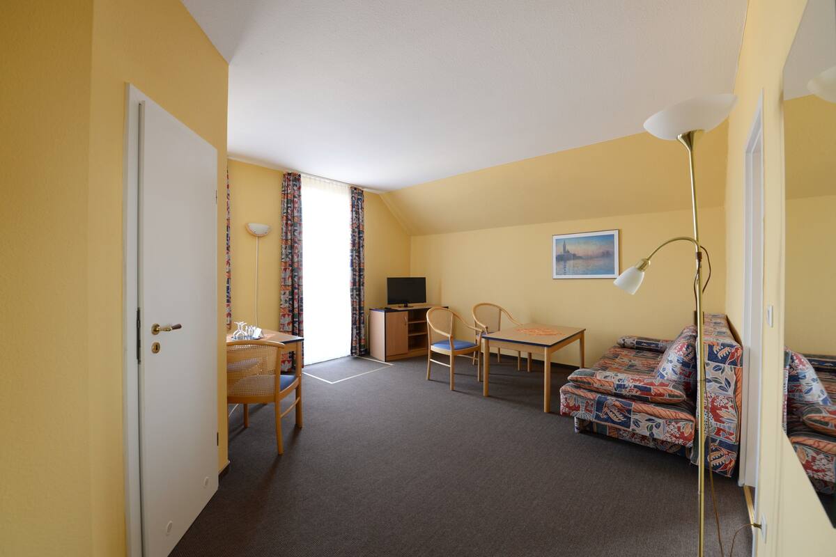 Una habitación con puerta, escritorio, silla, sofá, televisión y lámpara.