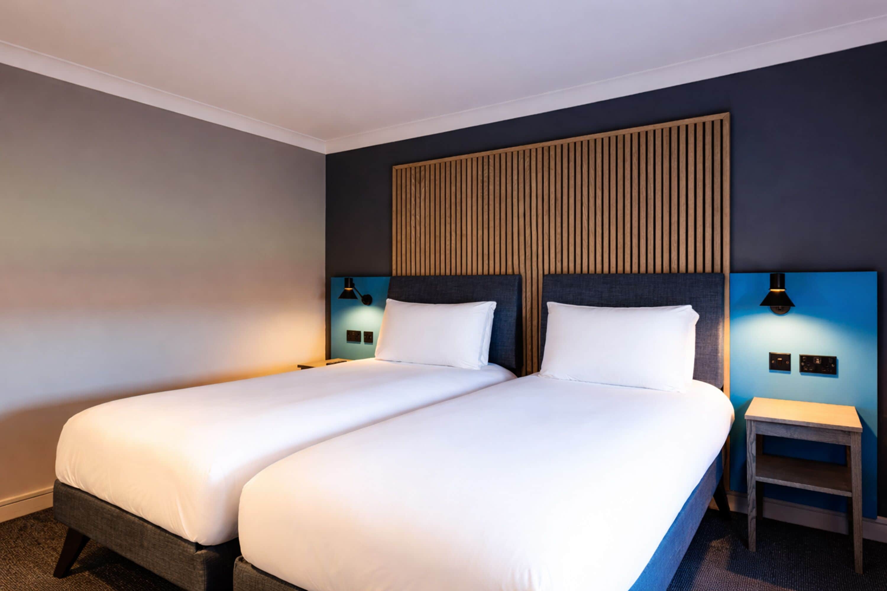 A hotel room with two beds, a wooden headboard, bedside lamps, and a small table.