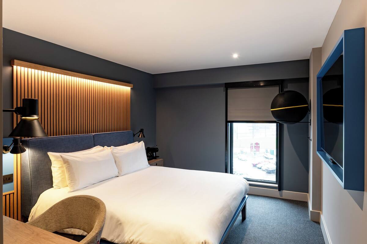 A modern hotel room with a large bed, a desk, a chair, a TV, and a window with a city view.
