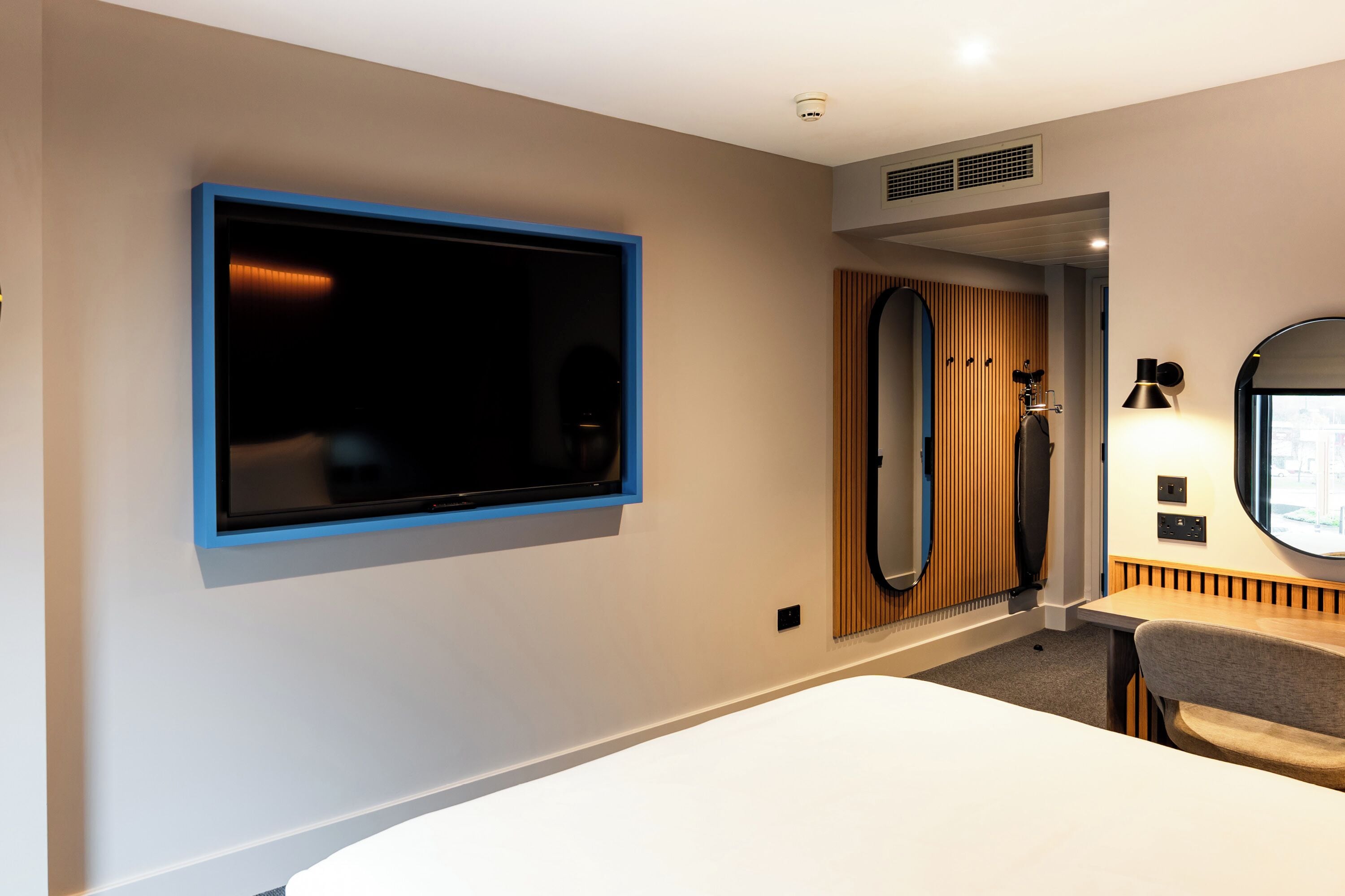 Ein modernes Hotelzimmer mit einem großen Flachbildfernseher an der Wand, einer Wandverkleidung aus Holzpaneelen, einem runden Spiegel und einem kleinen Tisch mit Lampe.