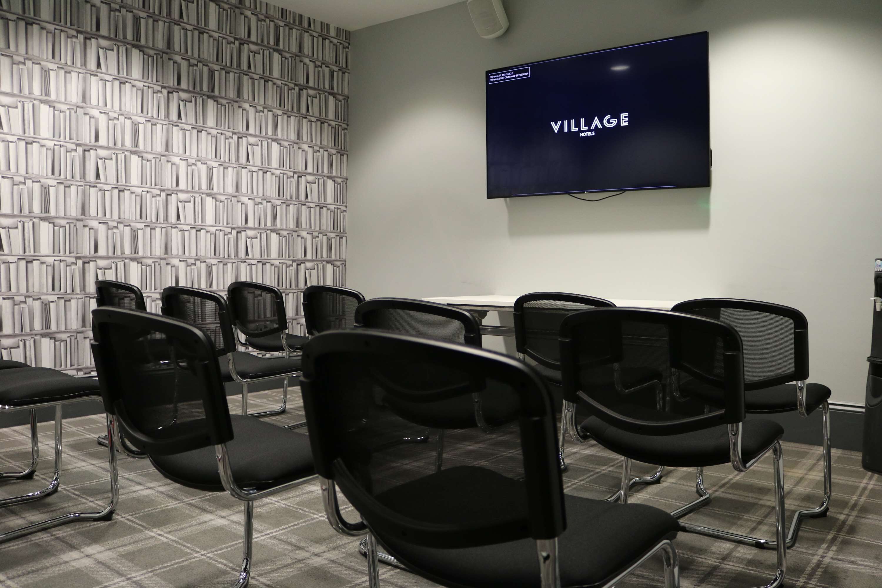 A conference room with a flat-screen TV displaying "VILLAGE HOTELS" on the wall, black chairs arranged in rows, and a patterned wall behind the chairs.