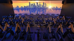 A gym with stationary bikes arranged in rows, a large screen displaying a sunset cityscape, and a wooden floor. - Village Hotel Solihull (Solihull)