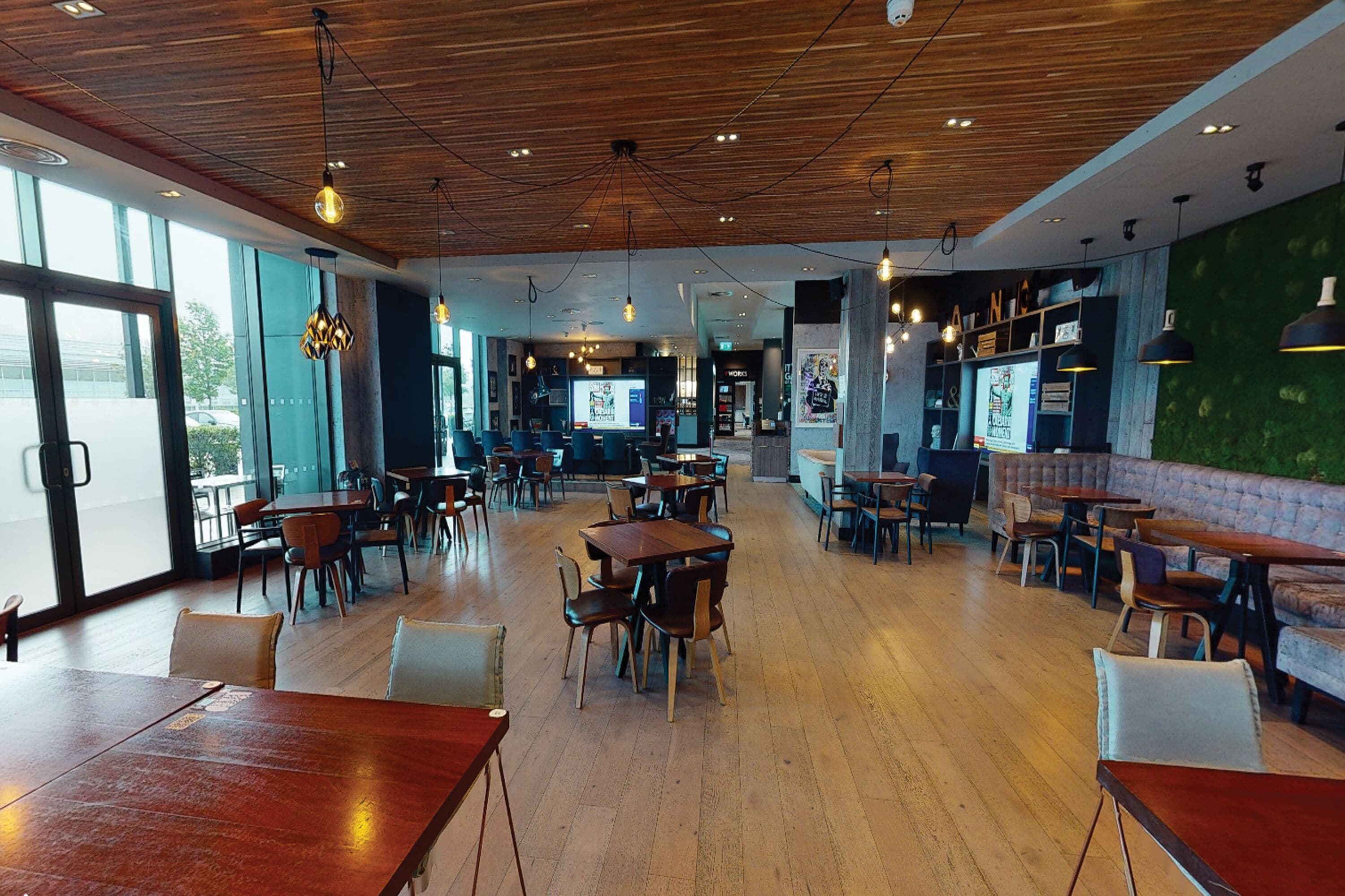 A spacious restaurant with wooden tables and chairs, a bar area, and a green wall with artwork.
