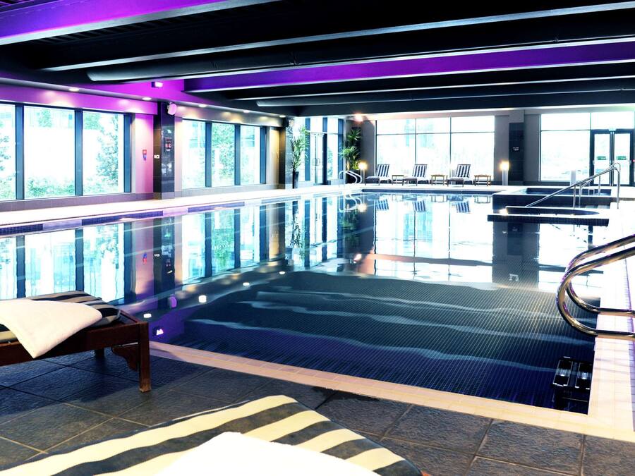 Una piscina interior con paredes de vidrio transparente, un área de descanso con cama y sillones, y vistas al exterior a través de amplios ventanales.