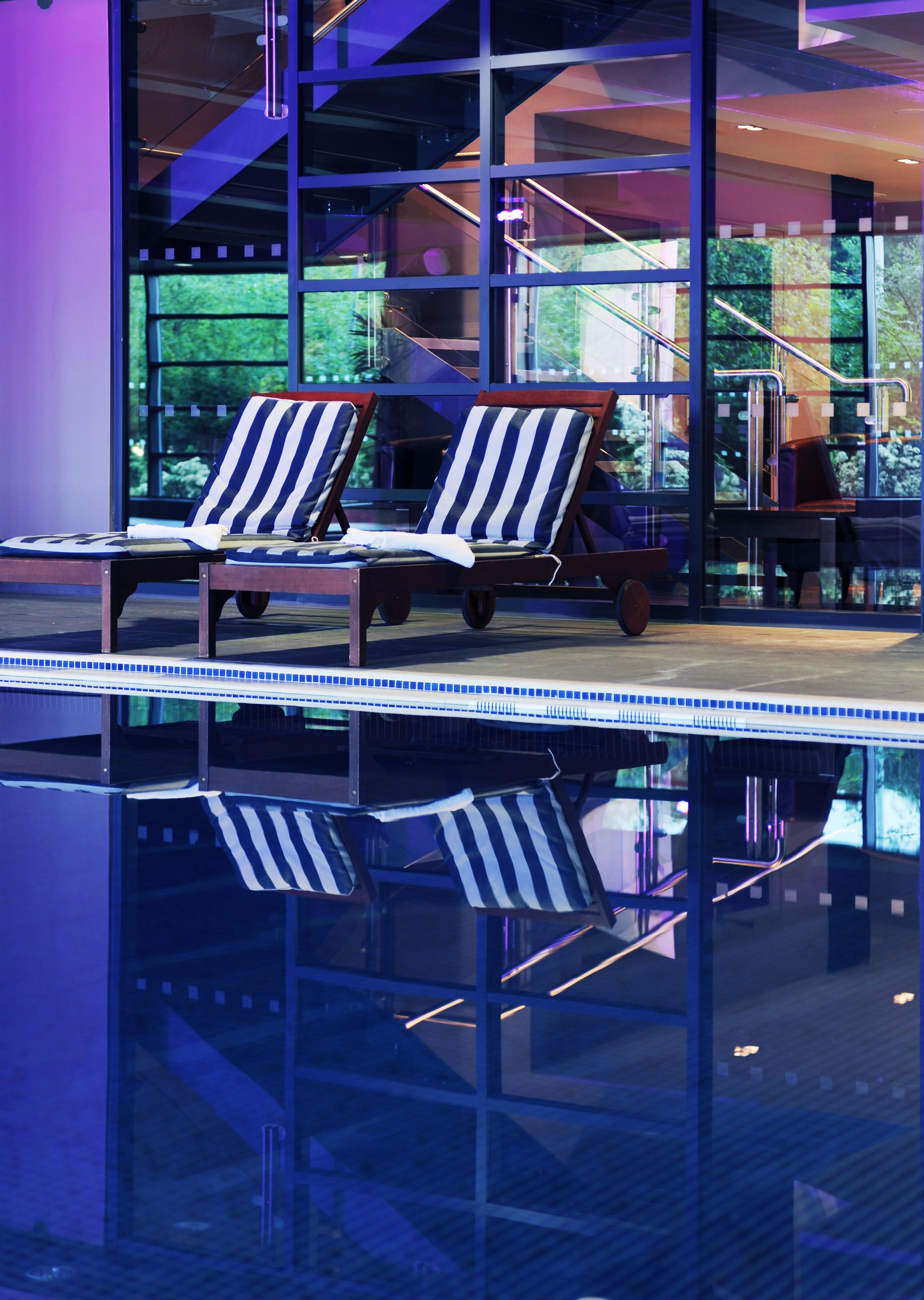 A modern indoor pool area with two lounge chairs and a glass wall reflecting the pool.