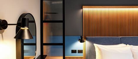 A modern hotel room with a bed, a desk, a lamp, and a wall-mounted control panel.