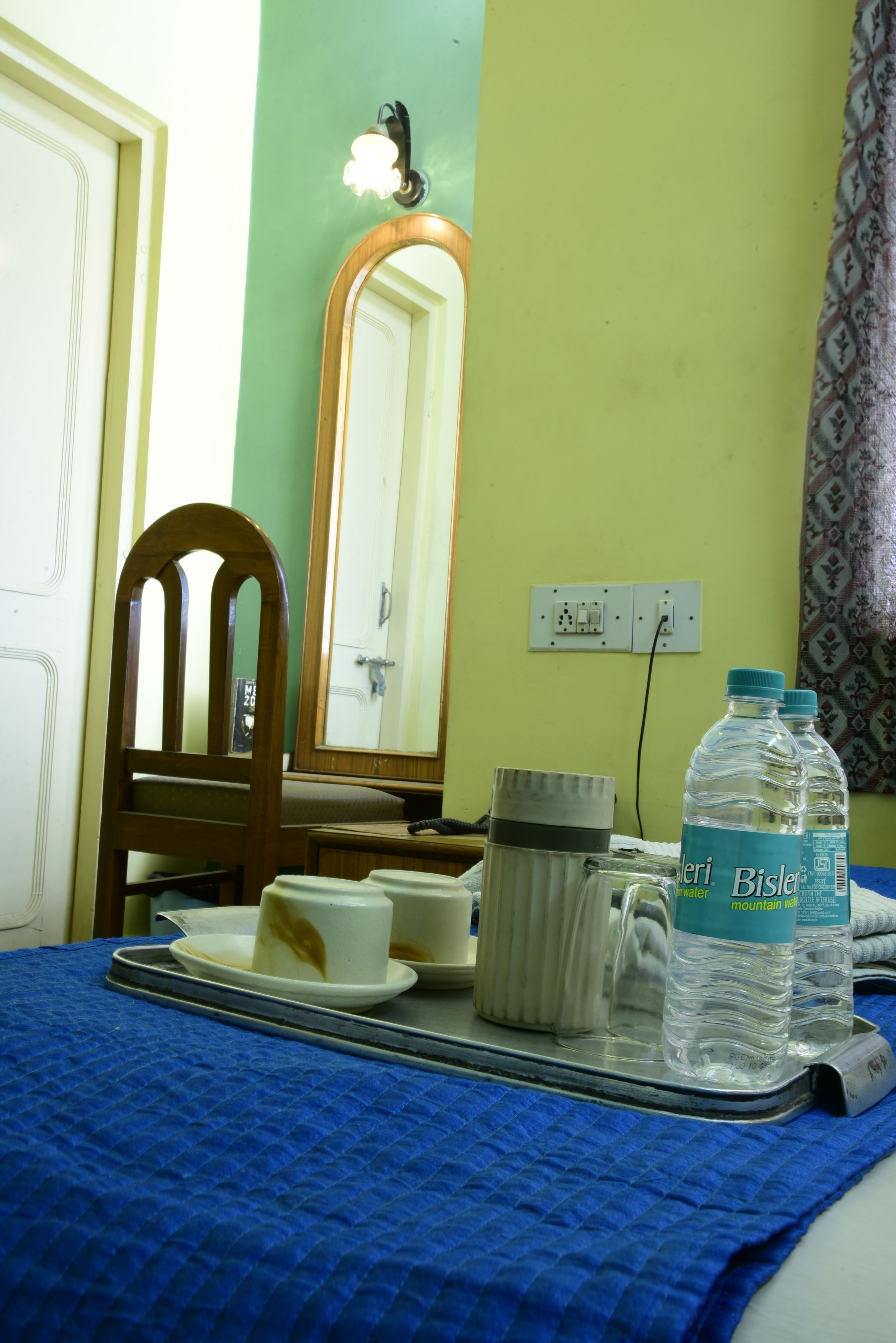 A hotel room with a bed, a tray with a cup and a bottle of water, a mirror, and a wall socket.