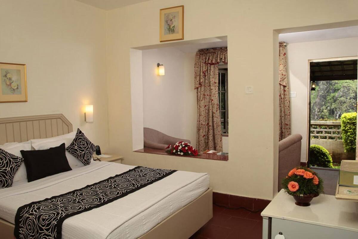 Ein Hotelzimmer mit einem Bett, einem Nachttisch mit Blumenvase, einem Fenster mit Vorhängen und Blick ins Grüne.