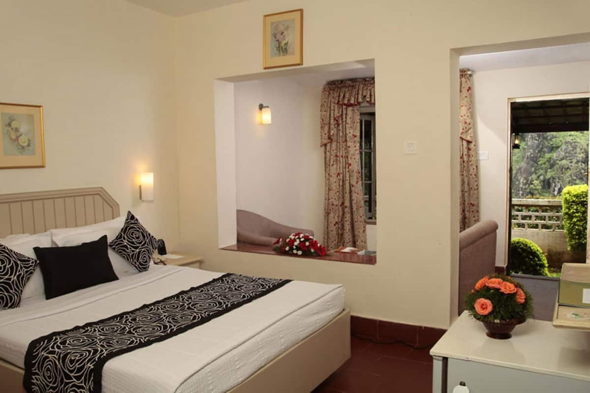 A hotel room with a bed, a bedside table with a vase of flowers, a window with curtains, and a view of greenery outside.