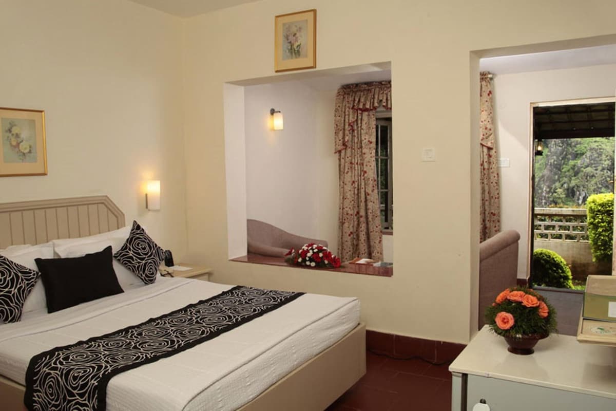 Habitación de hotel con una cama, una mesita de noche con un florero, una ventana con cortinas y vistas a zonas verdes.