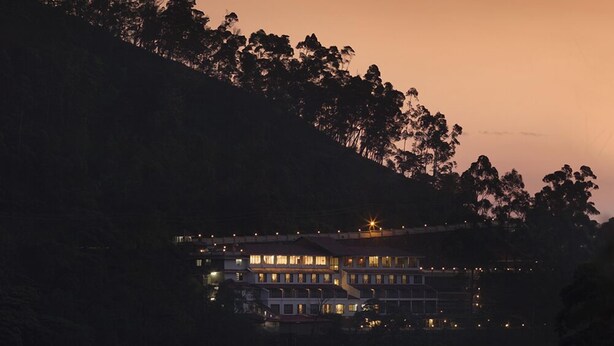 A multi-story building with multiple windows, surrounded by trees, during twilight. - Abad Copper Castle (Devikolam)