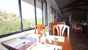 A dining area with a table set for two, large windows offering a view of a mountainous landscape, wooden chairs, and a view into the kitchen area. - Abad Copper Castle (Devikolam)
