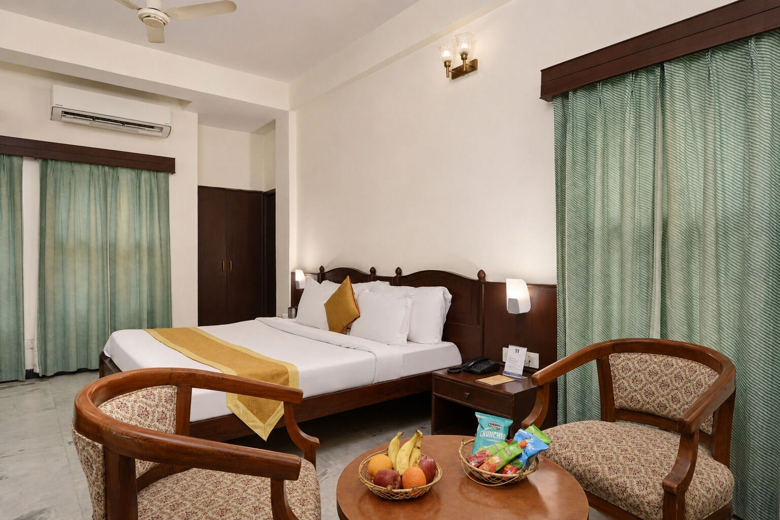 A hotel room with a bed, two chairs, a table with fruit and snacks, a bedside table with a phone, and curtains.