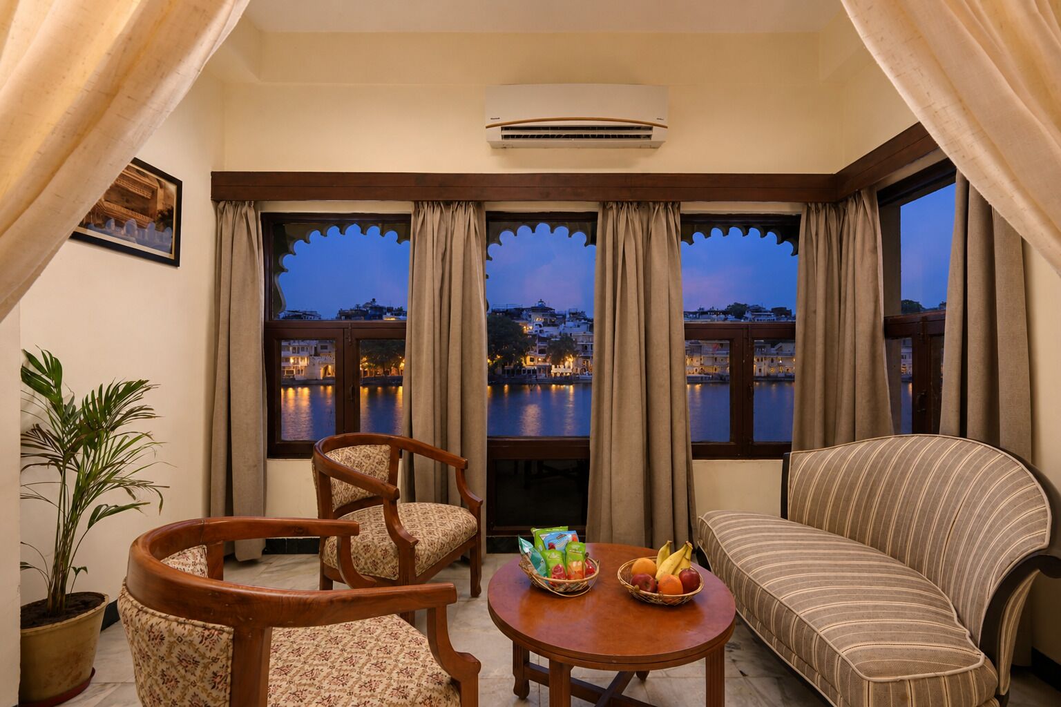 Habitación con vista a un muelle iluminado al anochecer, que incluye un sofá, dos sillas, una mesita con fruta y un equipo de aire acondicionado.