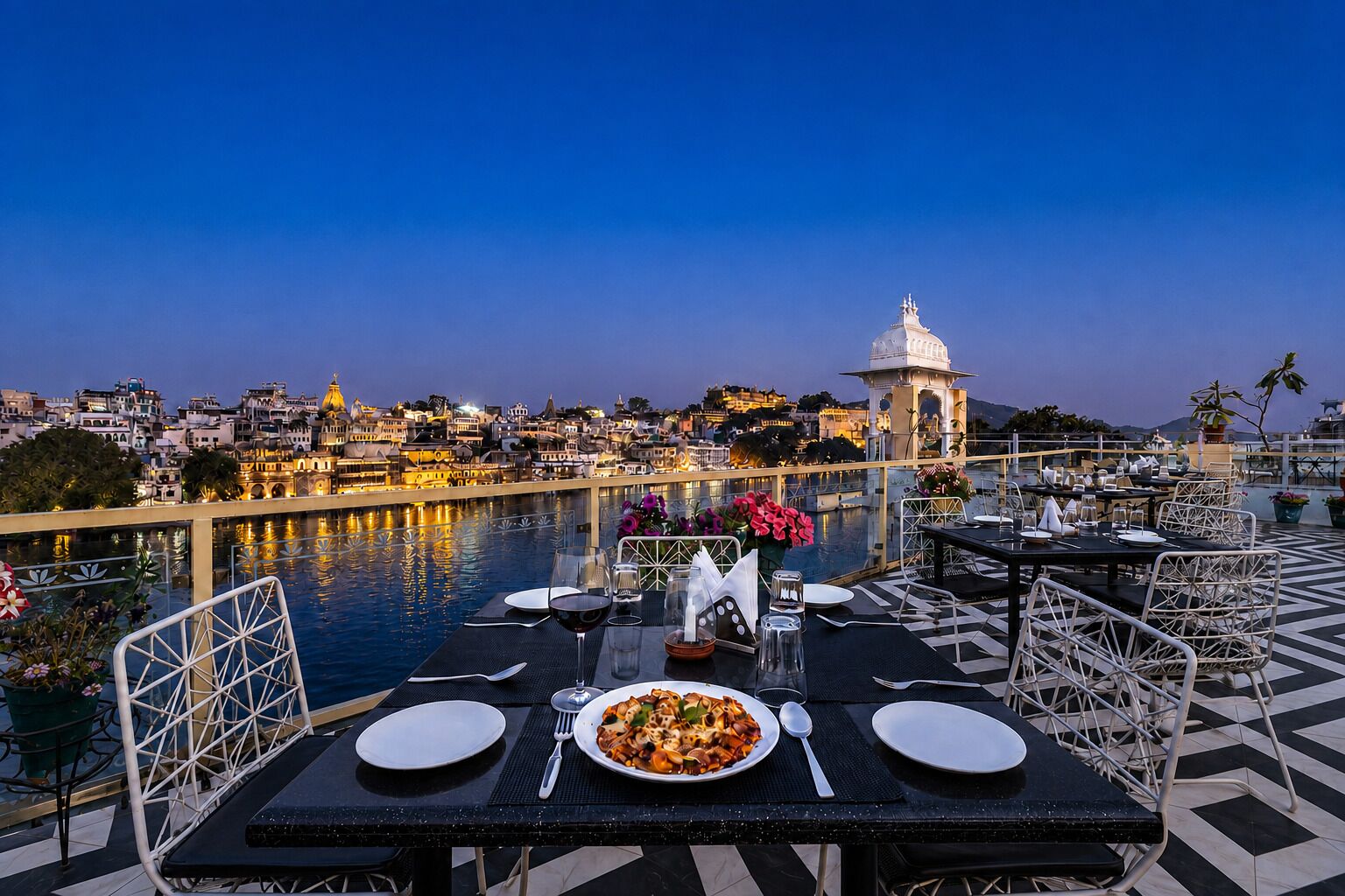 Una mesa de comedor con platos, cubiertos y una copa de vino tinto dispuesta para dos personas, con vistas a un cuerpo de agua y un paisaje urbano al fondo.