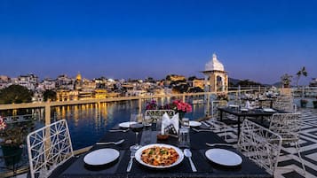 Una mesa de comedor con platos, cubiertos y una copa de vino tinto dispuesta para dos personas, con vistas a un cuerpo de agua y un paisaje urbano al fondo.