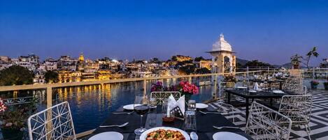 A dining table with plates, cutlery, and a glass of red wine set for two, overlooking a body of water with a cityscape in the background.