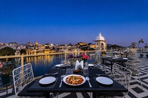 Una mesa de comedor con platos, cubiertos y una copa de vino tinto dispuesta para dos personas, con vistas a un cuerpo de agua y un paisaje urbano al fondo.