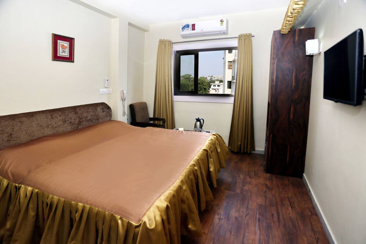 Habitación de hotel con una cama grande, una mesita pequeña, una silla, ventana con cortinas, equipo de aire acondicionado y un televisor en la pared.